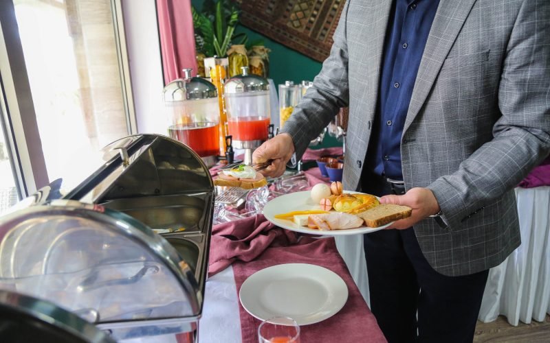side view man lays breakfast food egg sausages bun toast and cheese on a plate from the open buffet